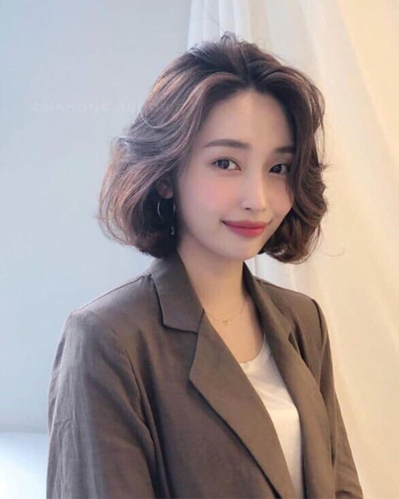 toc-ngan-uon-lon-to-1 Người phụ nữ tóc ngắn xoăn nhẹ, mặc áo vest nâu, áo thun trắng, đứng trước nền vải sáng.