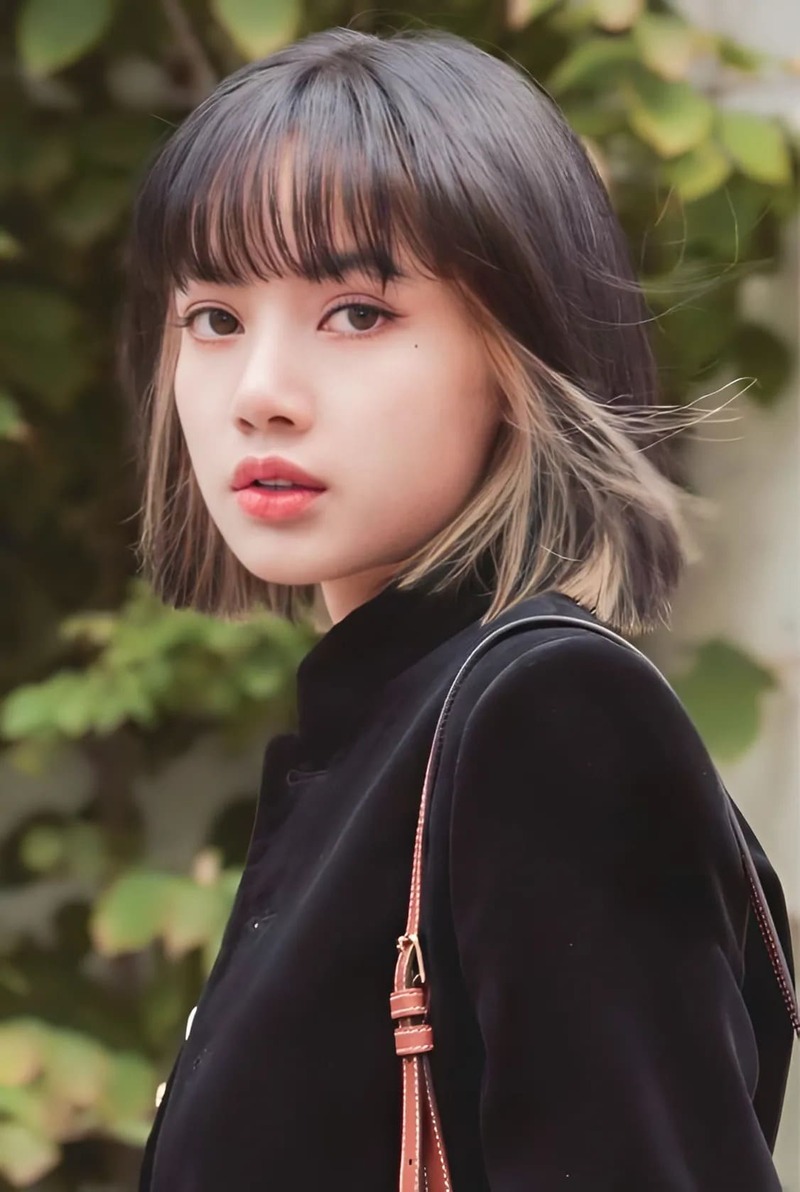 Người phụ nữ với tóc ngắn đứng ngoài trời, mặc áo đen, phía sau là nền lá cây xanh.