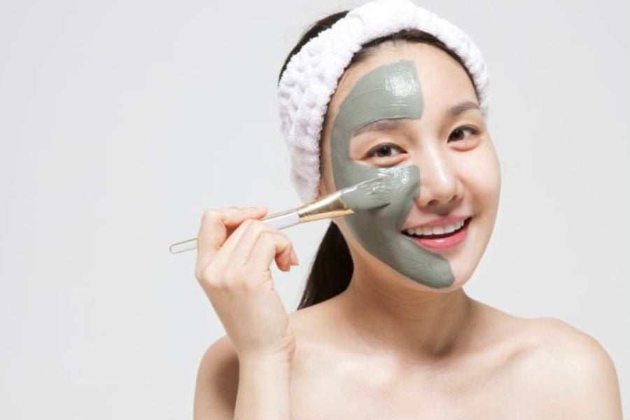 Người phụ nữ đang đắp mặt nạ bùn lên mặt, đội băng đô trắng, nền trắng.