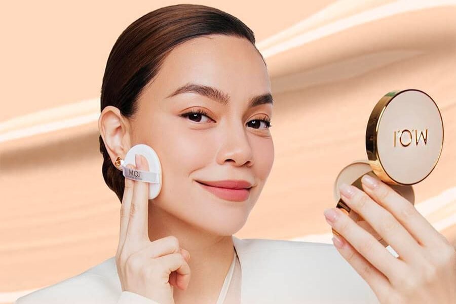 Người phụ nữ cầm gương và bông phấn, đang trang điểm trước nền màu be.