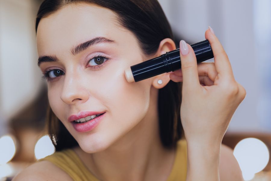 Người cầm cây son dưỡng môi, áp lên má, trong bối cảnh ánh sáng dịu nhẹ.