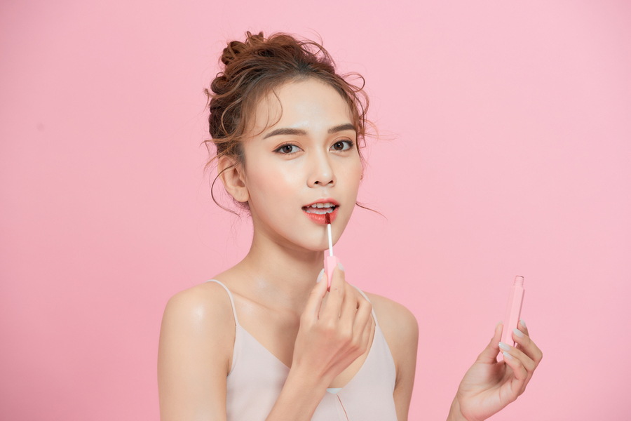 Người phụ nữ đang thoa son môi màu hồng, nền hồng, cầm lọ son trên tay.