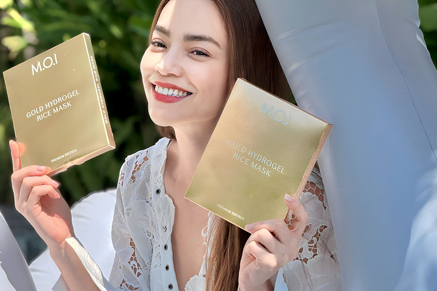 Người phụ nữ cầm hai hộp mặt nạ dưỡng da màu vàng, mặc áo trắng, ngồi trên ghế ngoài trời.