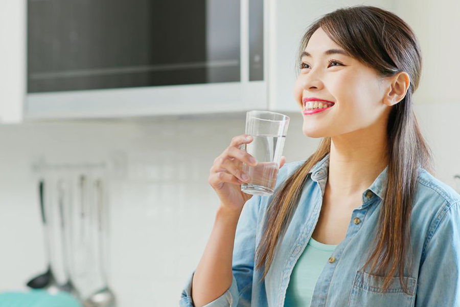 Người phụ nữ cầm ly nước, mỉm cười trong phòng bếp sáng sủa với lò vi sóng phía sau.