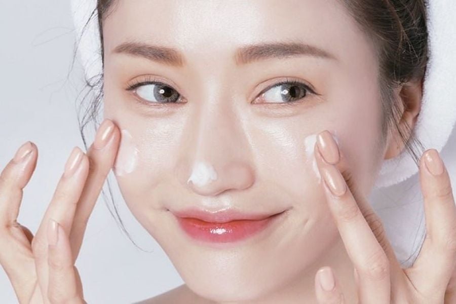 Người đang thoa kem dưỡng da lên má với nụ cười nhẹ, nền trắng.