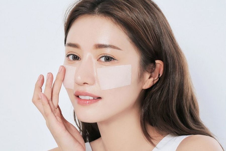 Người phụ nữ với miếng dán dưỡng da trên má, tay chạm nhẹ vào mặt, nền trắng.