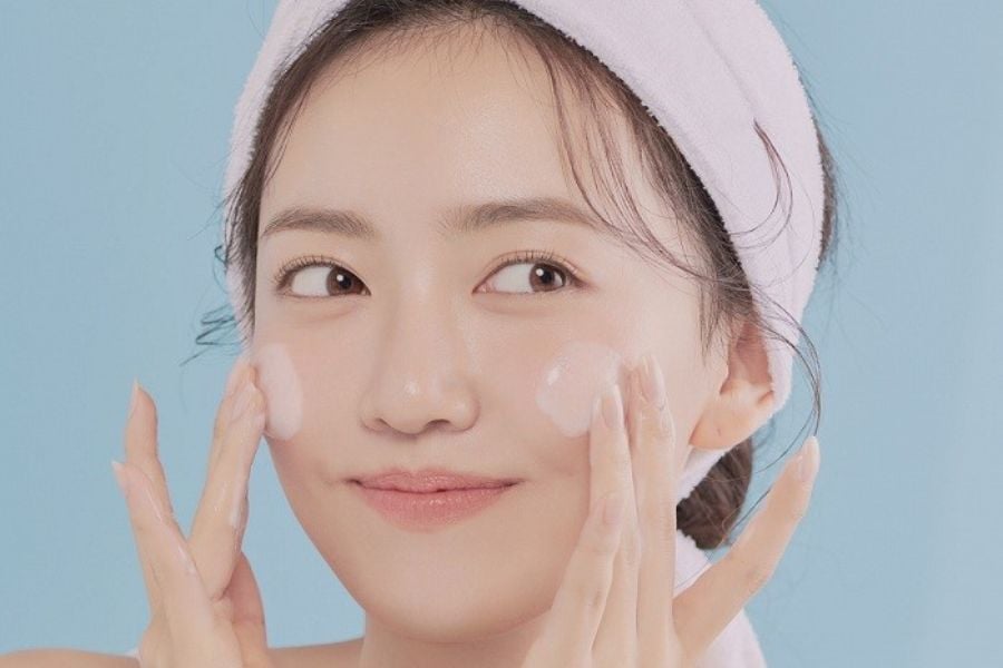 Người đang thoa kem dưỡng da lên má, đội băng đô màu hồng nhạt, nền xanh dương nhạt.