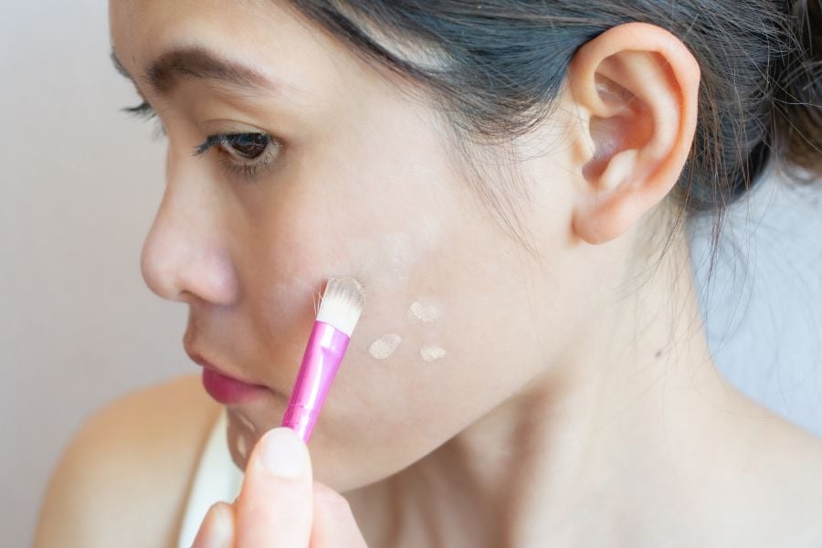Người đang dùng cọ trang điểm thoa kem nền lên má, phông nền đơn giản.