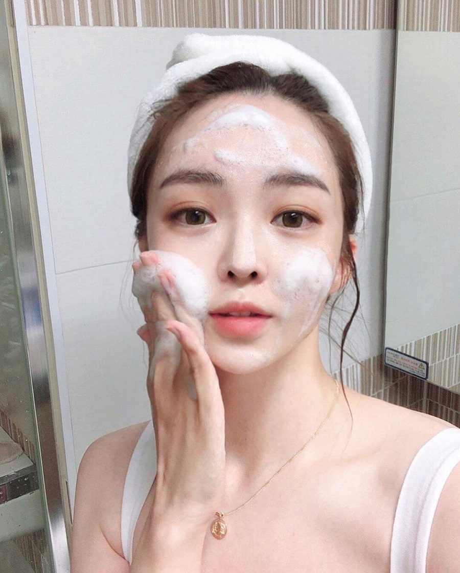 Người đang thoa sữa rửa mặt tạo bọt lên mặt trong phòng tắm, đầu quấn khăn trắng.