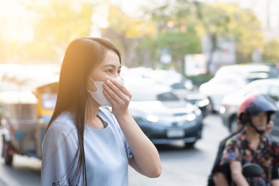Người phụ nữ đeo khẩu trang đứng trên vỉa hè, phía sau là đường phố tấp nập xe cộ.