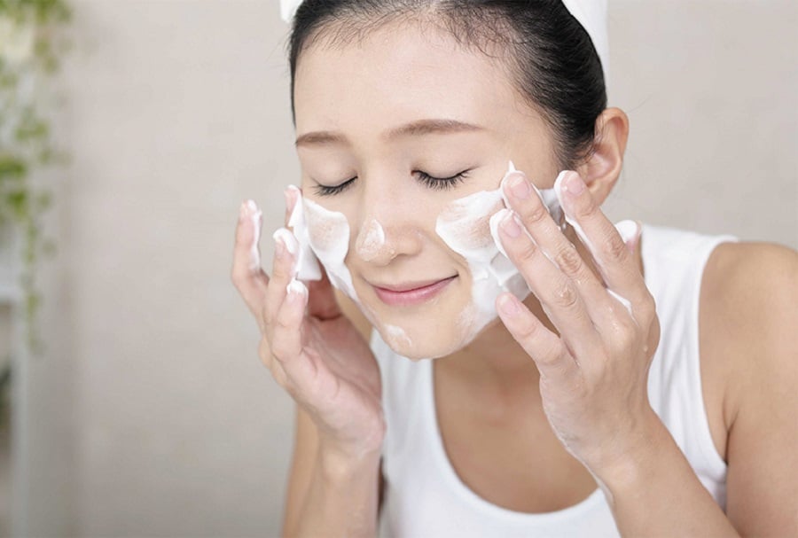 Người phụ nữ đang rửa mặt bằng sữa rửa mặt tạo bọt, mắt nhắm, vẻ mặt thư giãn.