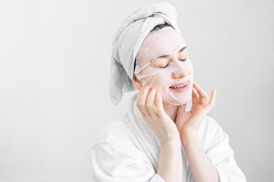 Người đang đắp mặt nạ dưỡng da, quấn khăn trên đầu, trong trang phục áo choàng trắng, nền trắng.