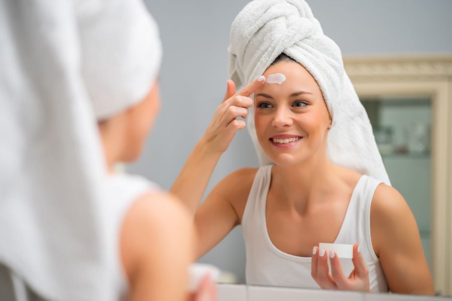 Người phụ nữ đứng trước gương, quấn khăn trên đầu và thoa kem dưỡng da lên trán, mỉm cười.