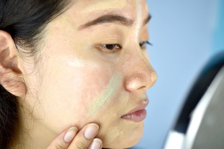 Người đang thoa kem dưỡng da lên má trước gương, nền xanh dương.