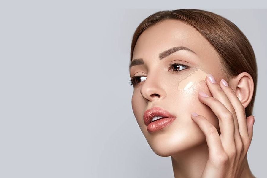 Người phụ nữ đang thoa kem nền lên má, nền xám, chú trọng vào chăm sóc da và trang điểm.