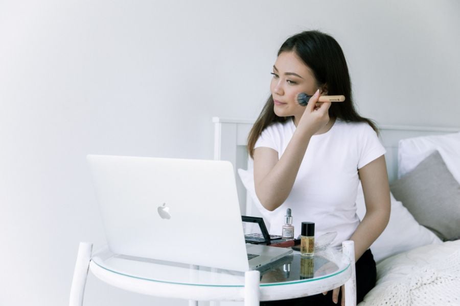 Người ngồi trên giường trang điểm, trước mặt là laptop, bàn có mỹ phẩm và cọ trang điểm.