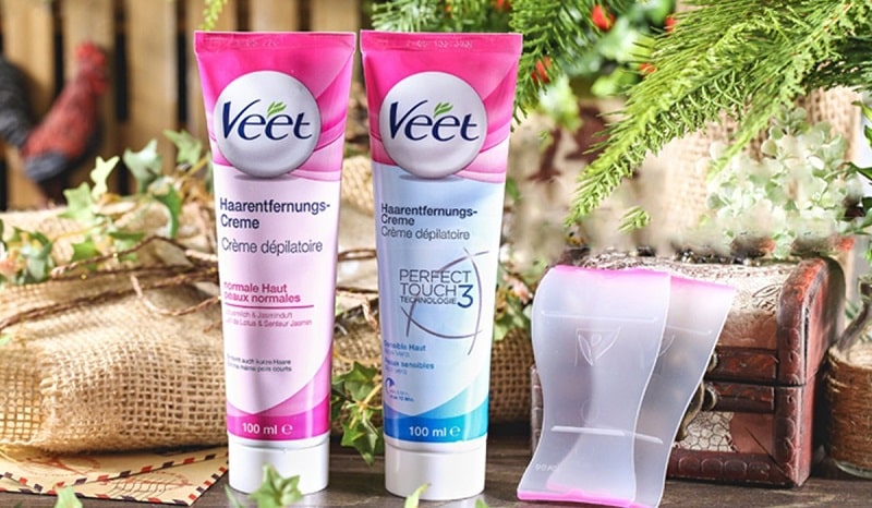 0f7f88e3-review-kem-tay-long-veet-1 Hai tuýp kem tẩy lông thương hiệu Veet cùng dụng cụ, đặt trên bàn với nền cây xanh và hộp gỗ.