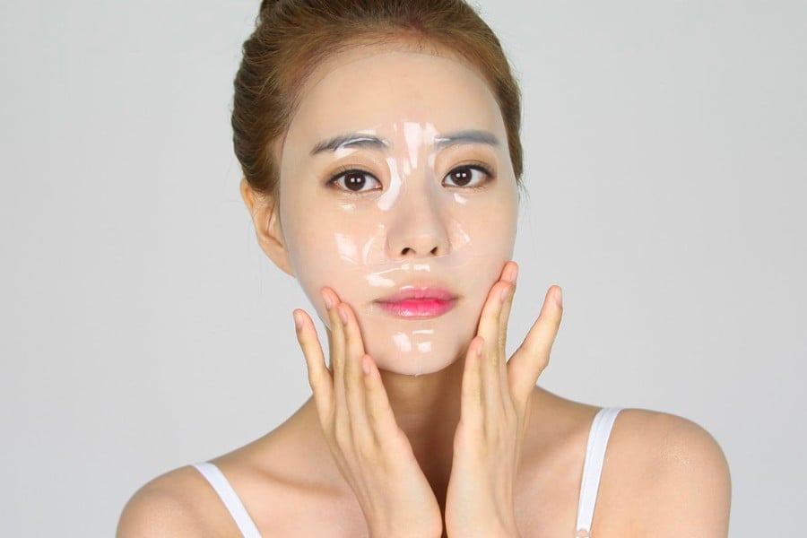 Người đang đắp mặt nạ dưỡng da trong suốt, tay chạm nhẹ lên mặt, nền xám nhạt.