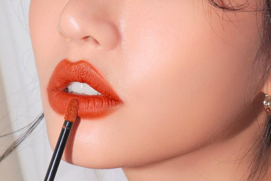 Cận cảnh một người đang tô son màu cam lên môi bằng cọ trang điểm.