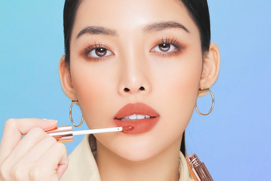 Người phụ nữ đang thoa son môi màu nâu đất, nền xanh, đeo khuyên tai vòng tròn.