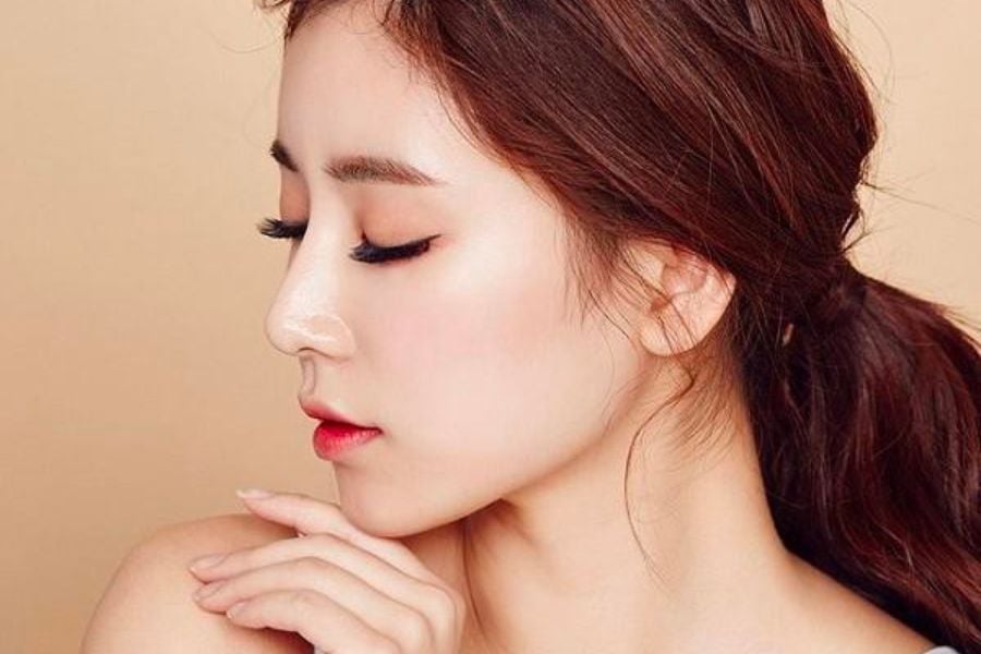 Người phụ nữ tóc nâu buộc gọn, trang điểm nhẹ, nhìn nghiêng, với nền màu be.