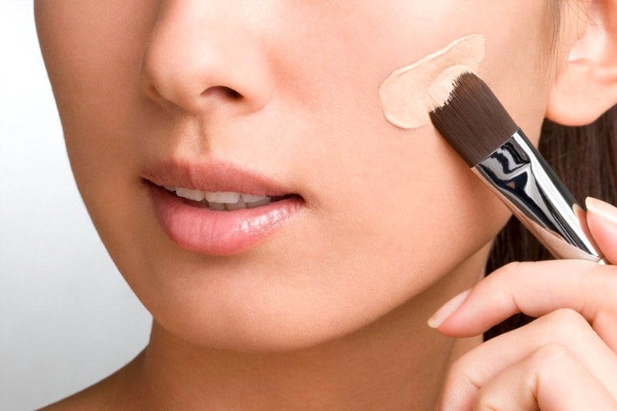 Người cầm cọ trang điểm thoa kem nền lên má, cận cảnh phần dưới khuôn mặt.