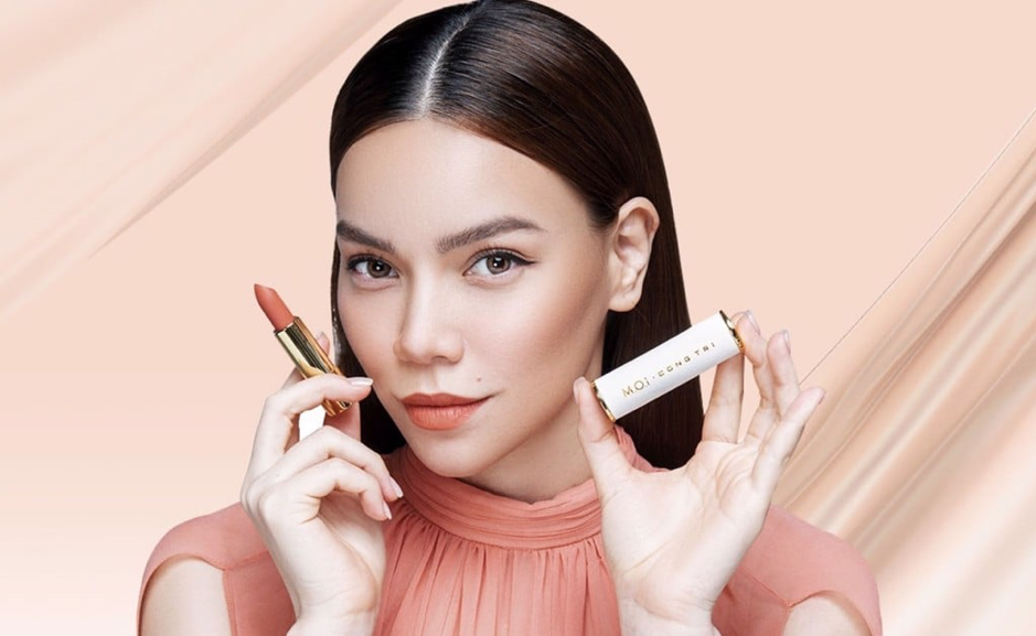 Người phụ nữ cầm son môi và hộp son, nền màu hồng nhạt, mặc áo màu hồng cam.
