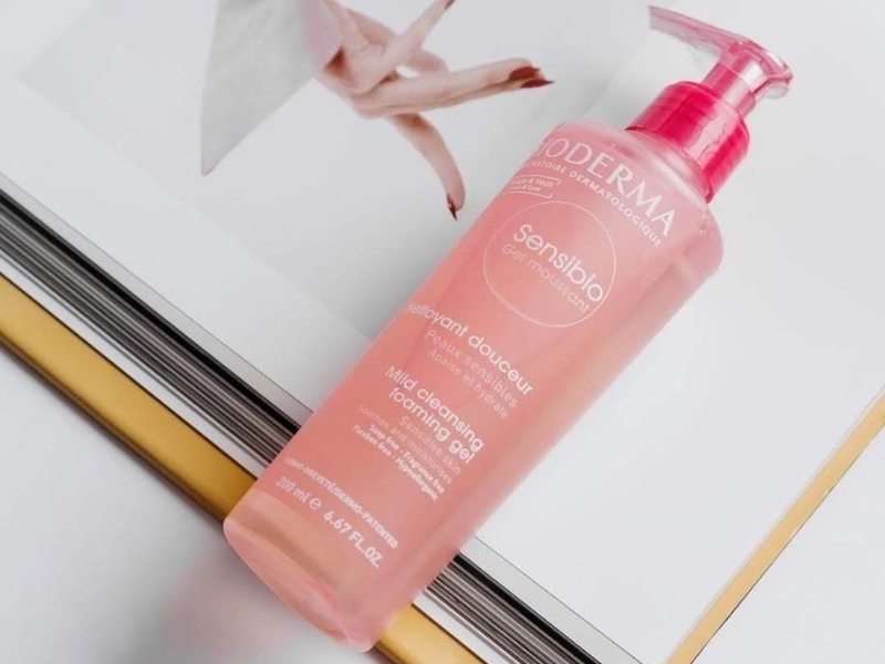1324b268-sua-rua-mat-cho-da-dau-03 Chai gel rửa mặt màu hồng đặt trên một cuốn sách mở, nắp chai có vòi bơm màu hồng.