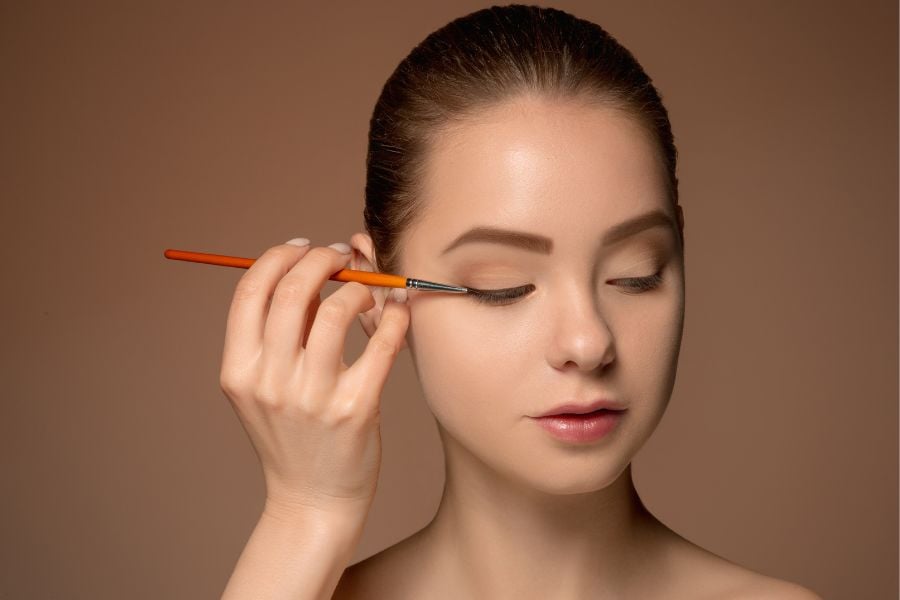 Người phụ nữ đang trang điểm mắt bằng cọ, nền màu nâu, đôi mắt nhắm hờ, biểu cảm tập trung.