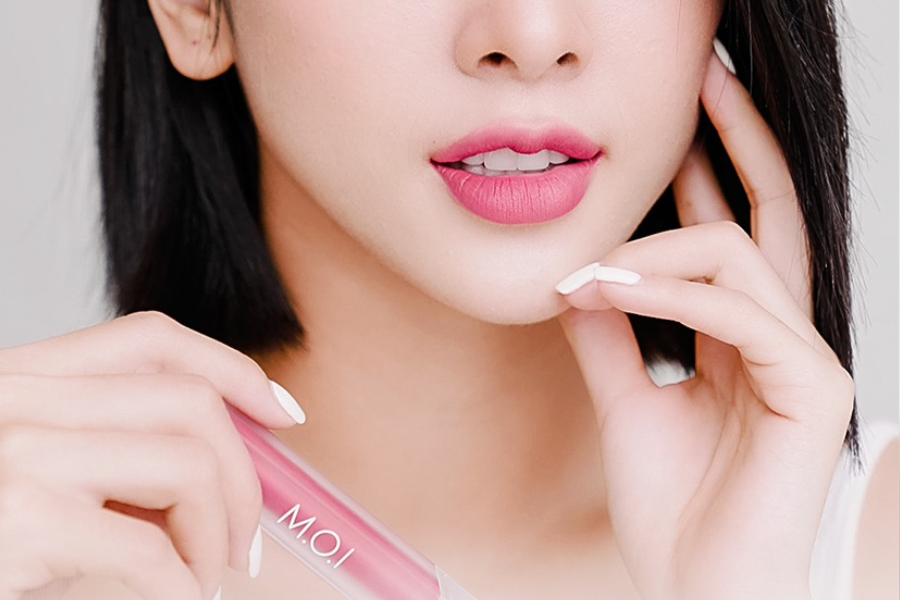 Người cầm thỏi son môi màu hồng, môi được tô son hồng, tay chạm nhẹ lên má, nền trắng.