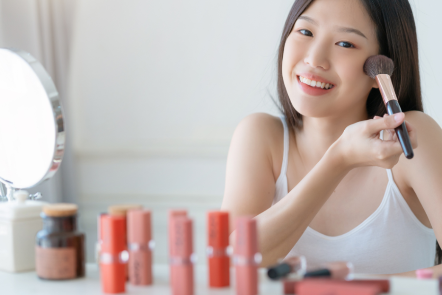 Người phụ nữ cầm cọ trang điểm, mỉm cười trước gương, xung quanh là các sản phẩm trang điểm khác.