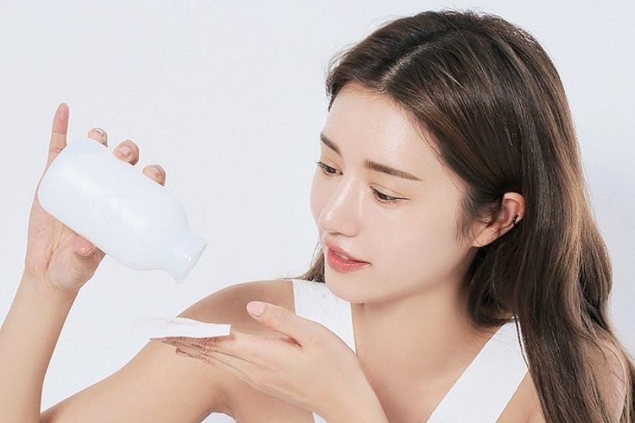 Người phụ nữ đang đổ dung dịch từ chai nhựa trắng lên miếng bông để chăm sóc da mặt.
