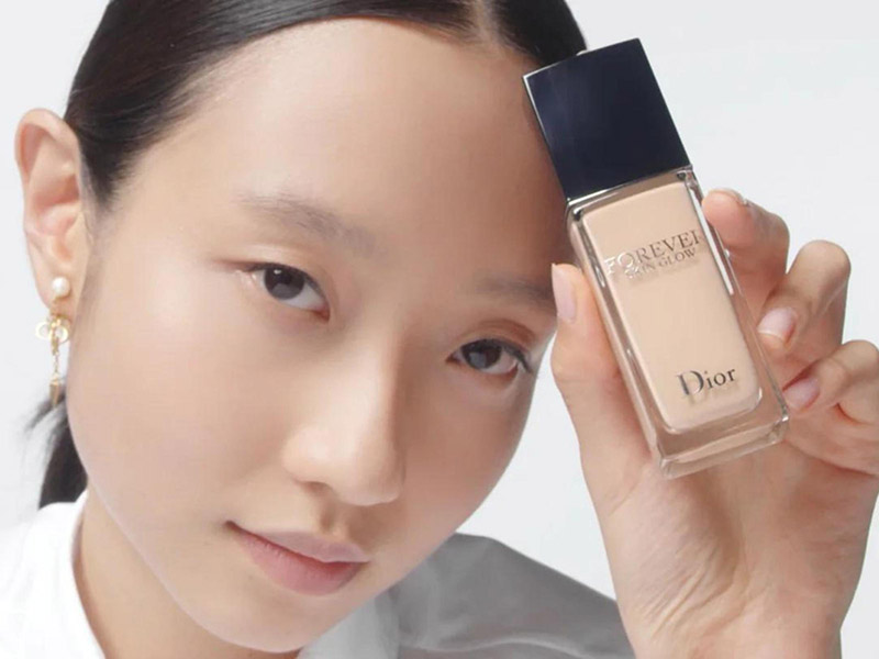 2d067849-kem-nen-cho-da-kho-9 Người cầm chai kem nền Dior, mặt nghiêng và biểu cảm nhẹ nhàng, nền trắng đơn giản.