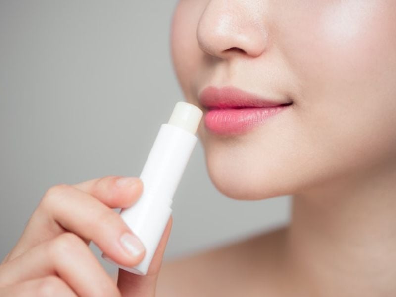 Người đang thoa son dưỡng môi không màu lên môi, cận cảnh khuôn mặt và tay cầm thỏi son.
