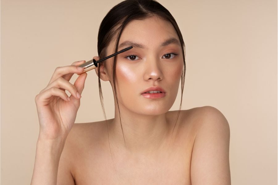 Người phụ nữ đang chải lông mày bằng cọ mascara, nền màu be, trang điểm nhẹ nhàng, tóc buộc gọn.