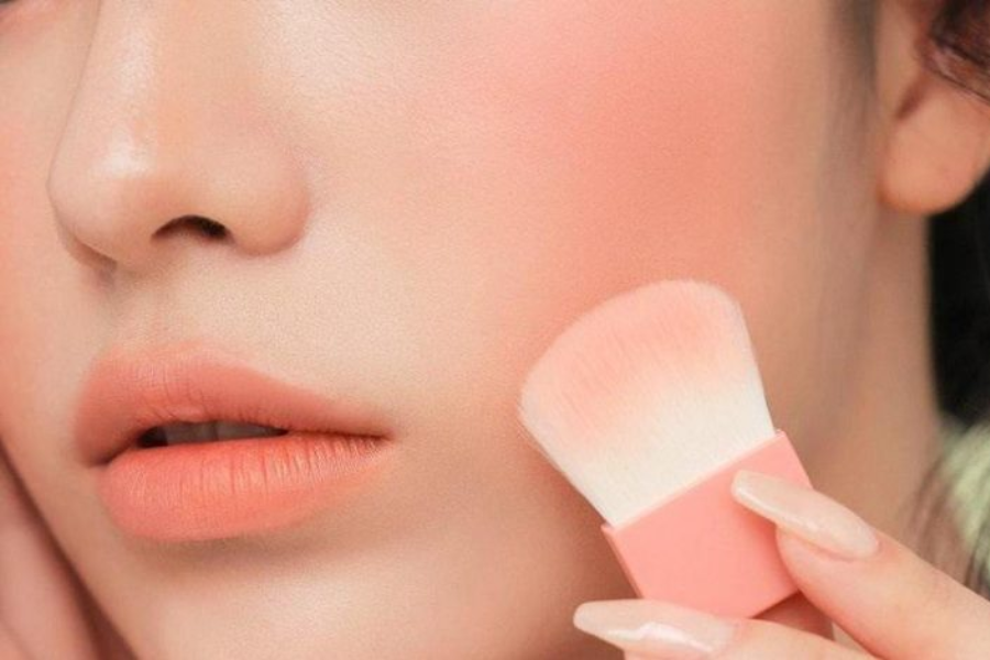 Cận cảnh một người đang dùng cọ trang điểm để thoa phấn má hồng lên gò má.