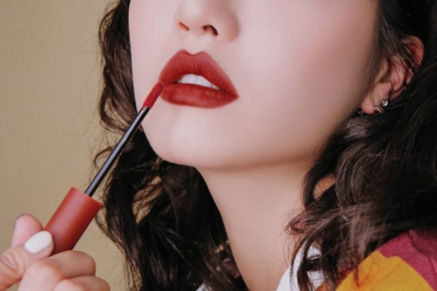Người cầm thỏi son đỏ, tô lên môi, với tóc xoăn nhẹ và nền màu be.
