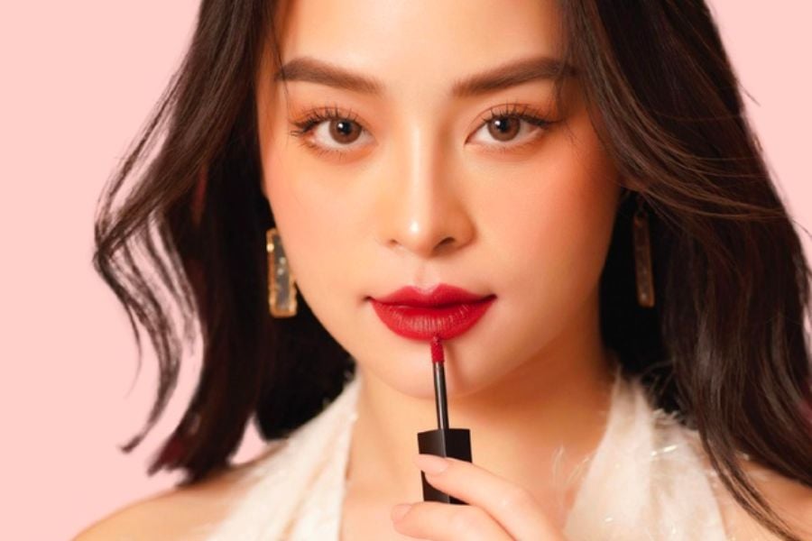 Người phụ nữ thoa son đỏ lên môi, nền màu hồng nhạt, đeo khuyên tai dài và mặc áo sáng màu.