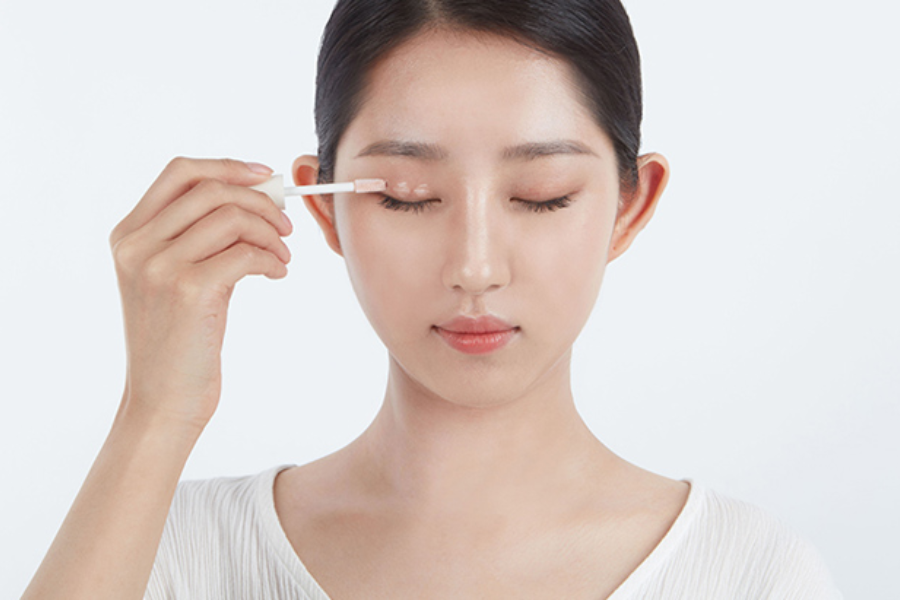 Người phụ nữ đang thoa sản phẩm trang điểm lên mí mắt bằng cọ, mắt nhắm, nền trắng.