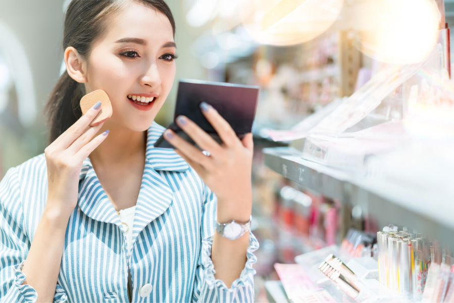Người phụ nữ cầm gương nhỏ và miếng trang điểm, đứng trước kệ mỹ phẩm trong cửa hàng.