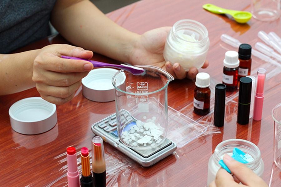 Người đang pha trộn nguyên liệu trong cốc thủy tinh trên bàn gỗ, xung quanh có nhiều chai lọ và dụng cụ.