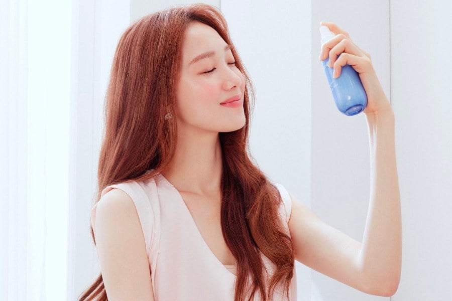 Người phụ nữ đang dùng chai xịt dưỡng da màu xanh, mắt nhắm, tóc dài, mặc áo không tay màu nhạt.