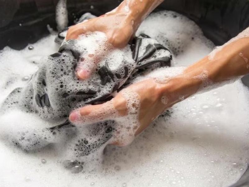 Hai tay đang giặt quần áo màu đen trong chậu nước xà phòng, tạo nhiều bọt trắng.