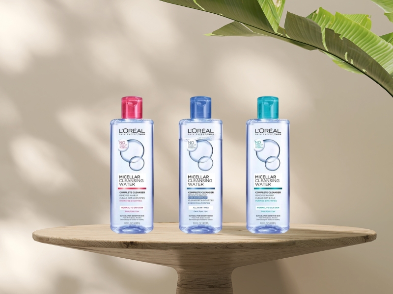 3ac3a19f-nuoc-tay-trang-loreal-cho-da-nhay-cam-1 Ba chai nước tẩy trang đặt trên bàn gỗ, nền màu nâu, có cây xanh trang trí bên phải.