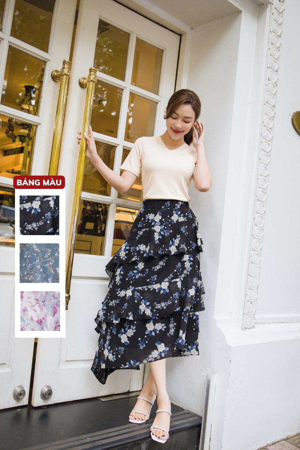 10 Cách Phối Đồ Với Chân Váy Hoa Nhí Giúp Bạn Tỏa Sáng Mùa Hè 8 Người phụ nữ mặc áo trắng và váy hoa tối màu đứng trước cửa, bên cạnh là bảng mẫu vải.