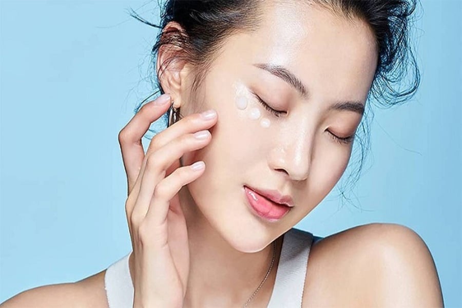 Người phụ nữ thoa kem dưỡng da lên mặt, nền xanh, mắt nhắm và vẻ mặt thư giãn.