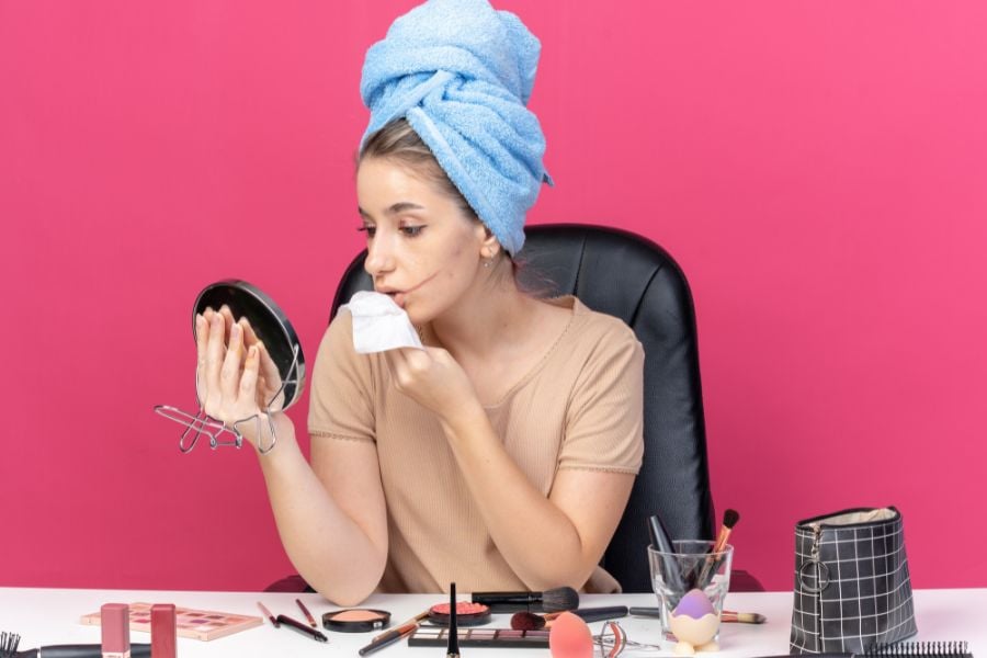 Người phụ nữ ngồi trước bàn trang điểm, lau mặt bằng khăn giấy, đầu quấn khăn màu xanh, nền hồng.