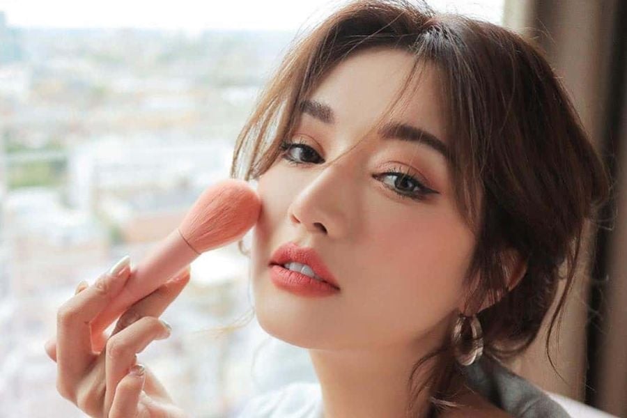 Người phụ nữ trang điểm, dùng cọ má hồng, nền cảnh mờ của thành phố qua cửa sổ.