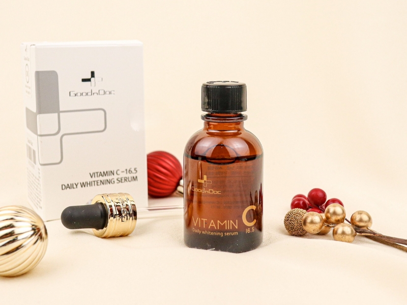Chai serum vitamin C và hộp đựng trên nền trắng, trang trí với quả cầu và nhánh cây.