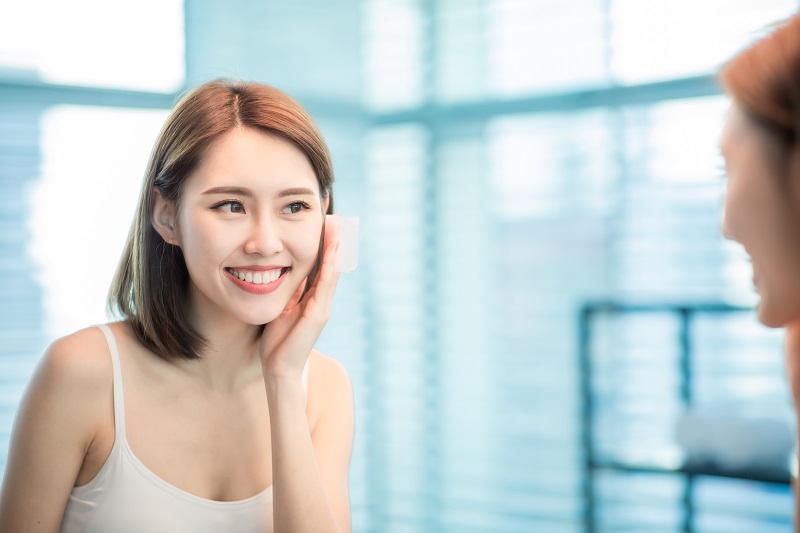Người phụ nữ đang sử dụng bông tẩy trang trên mặt, đứng trước gương trong phòng sáng sủa.